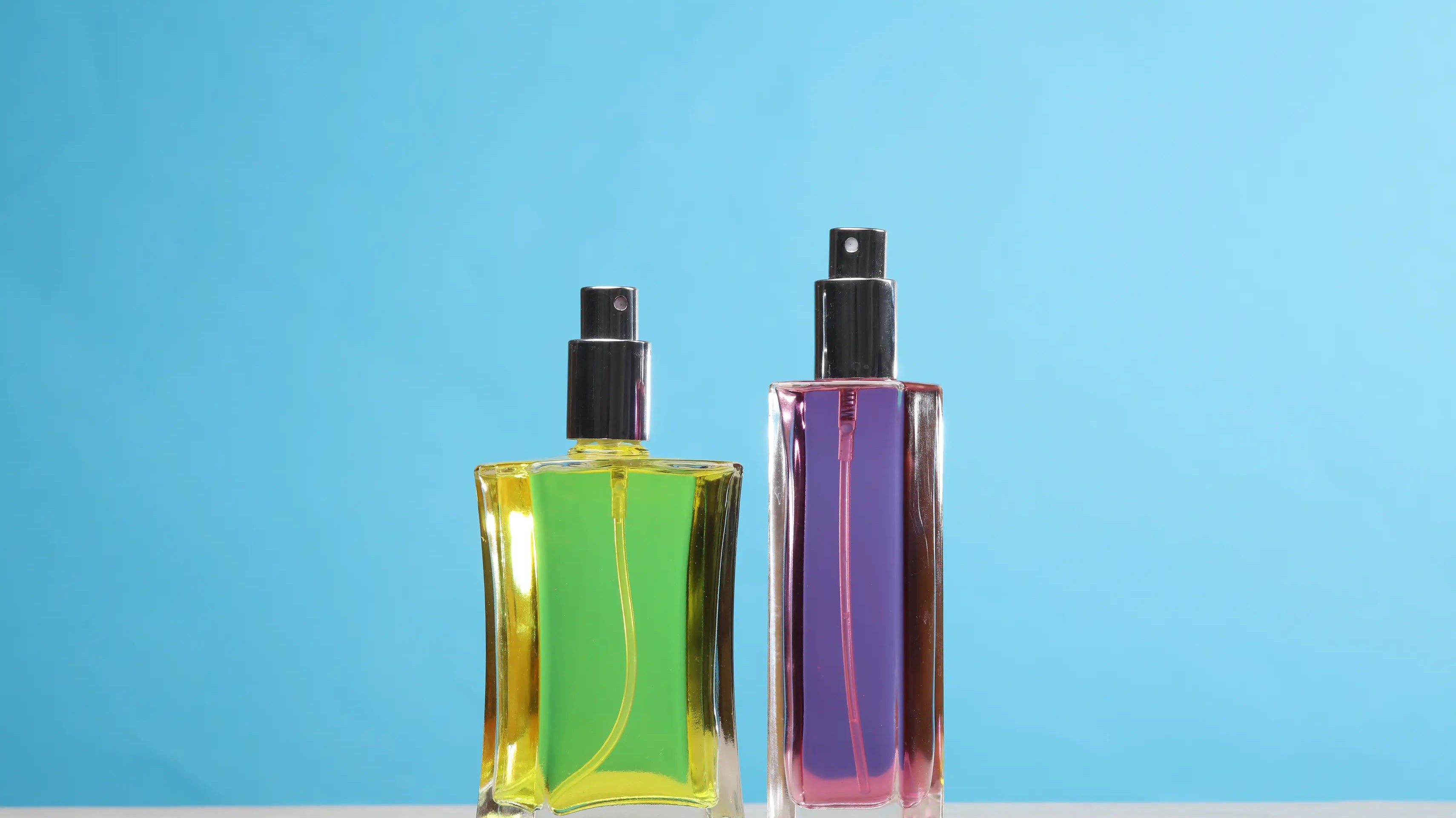 Two perfume bottles in different shapes and colors set on a blue backdrop.