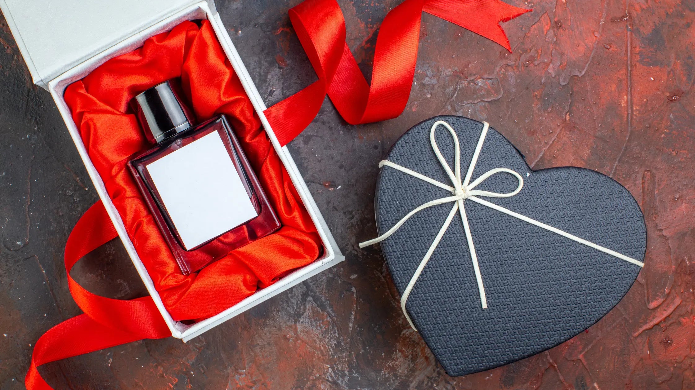 A Valentine's day heart-shaped gift box and a perfume bottle in a white gift box, placed atop a reddish-dark background.