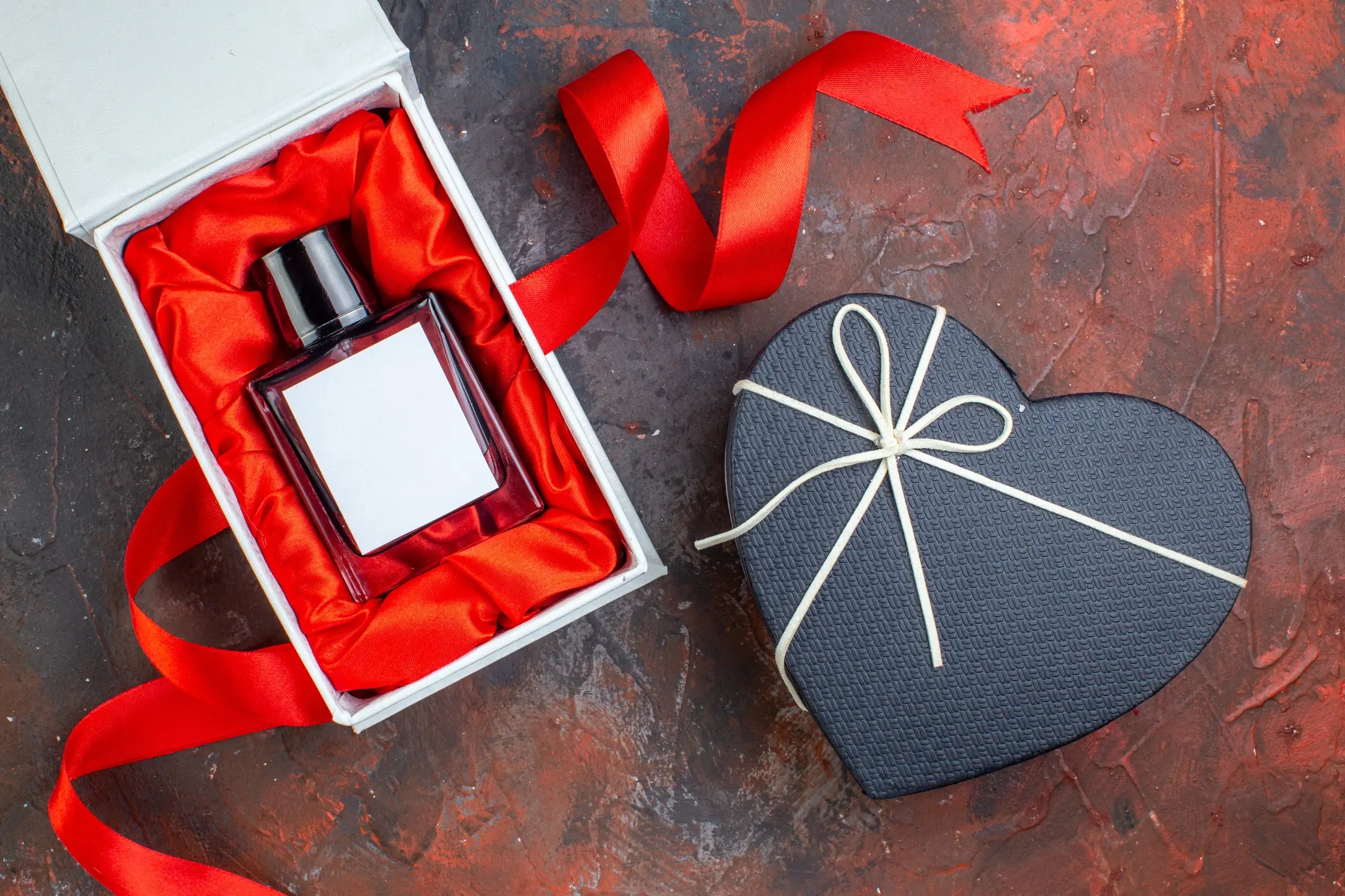 A Valentine's day heart-shaped gift box and a perfume bottle in a white gift box, placed atop a reddish-dark background.
