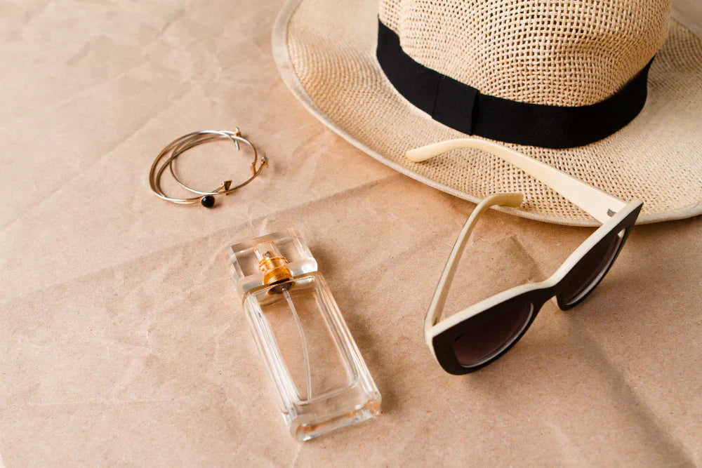 A straw hat, sunglasses, bracelets and a perfume bottle on a plain brown paper background. 